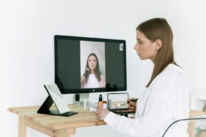 A woman applies makeup while following an online beauty tutorial on a desktop screen.